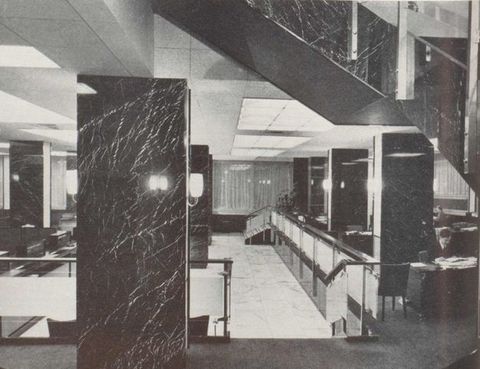 Black and white photograph. An interior view of a modern building, looking through the lobby area to curtained windows beyond. Part of an angular staircase can be seen on the right. Below the staircase, several desks and tables can be seen. A man sits behind one, reading from a stack of paper spread out before him.