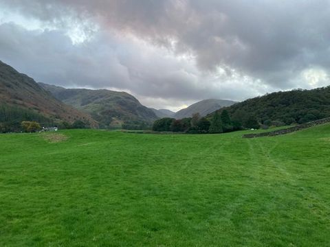 Colour photograph. A document of a landscape scene, with a large expanse of flat, green grass in the foreground, that leads towards distant mountains and a billowing sky. A partial stone wall can be seen on the right, and a small white and grey house is just visible on the left.
