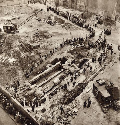 Black and white photograph. An aerial view looking over the construction site of an archaeological dig, which is filled with visitors. A roped-off area of ground has been dug into, and is surrounded by people looking into it. Several large-scale construction tools can be seen in the space beyond, along with queues of more visitors. 