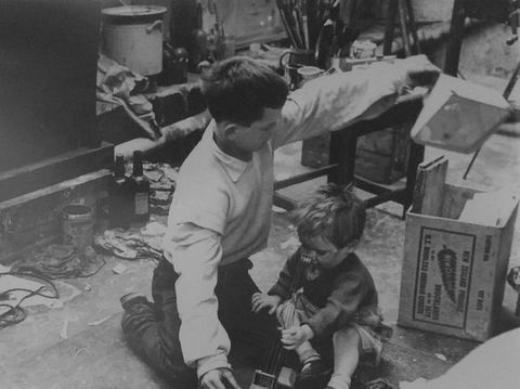 Black and white photograph. Two young children kneel on the floor of an artist’s studio, surrounded by materials including rags, boxes, paintbrushes and old cans. The younger of the two boys appears to play with a toy guitar, while the elder reaches over his head to pull out the contents of a wooden box. 