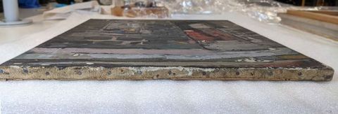 Colour photograph. The view of a painting laid out onto a table, seen from a side-on view. A row of nails holding the canvas to the frame can be seen. In the distance, art packing materials are just visible. On the right, part of another artwork or frame can be seen.
