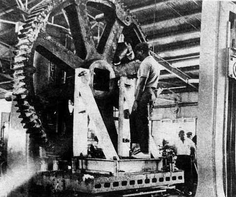 Black and white photograph. A view into the interior of a factory, where people carry out work. The image is dominated by a large, serrated wheel held in place by a frame. A black person is seen from behind facing the wheel, seemingly captured mid-work. 