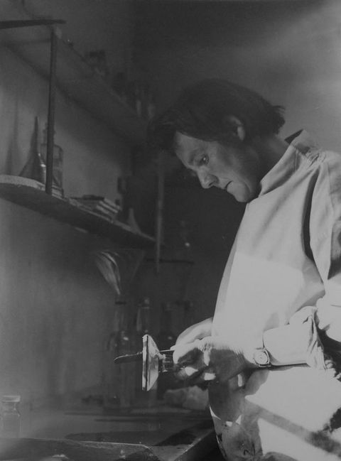 Black and white photograph. A document of a person leaning over a worktop while scraping together two different tools. They are surrounded by a variety of bottles, jars, and containers on the worktop and shelves around them.
