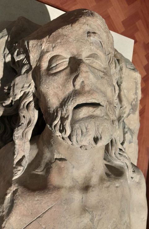 Colour photograph. The close-up view of a stone statue, showing a man’s head seen from above. He has a beard and long curly hair, with eyes closed and lips slightly parted. The top part of his head on the left has broken away. Behind his head a white plinth and parquet flooring is just visible.