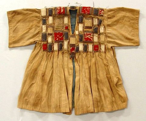 Colour photograph. Documentation of a gold-coloured top with short, wide sleeves, a V-neck, and a loose, wide bodice with an open slit at the front. Across the upper breast a series of vividly coloured leather squares and rectangles have been stitched into the fabric to create a striking, textural pattern.