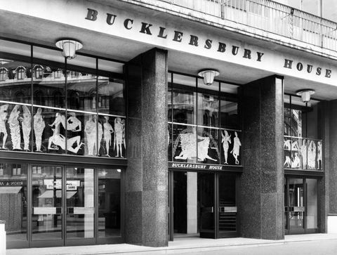 Black and white photograph. The front façade of a building is visible, with sliding glass doors at ground level, and glass windows on the first level. Above the main entrance reads the sign ‘Bucklersbury House’. A series of cut-out drawings of people have been pasted to the first-floor windows, some standing, and others reclining.