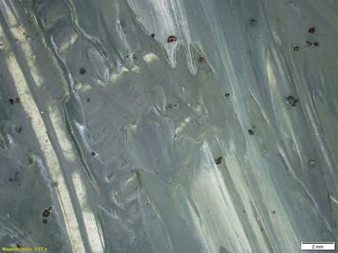 Colour photograph. Close-up detail of a passage of blue-grey paint with a textural surface. A scraped away area runs diagonally across the left side. On the lower left yellow text reads, ‘Magnification: 0.67 x’. On the right, a small white rectangle bears text reading ‘2 mm’.