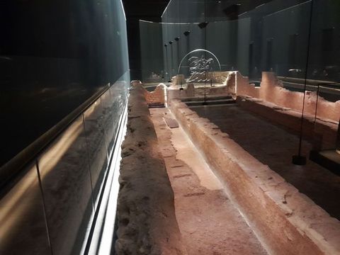 Colour photograph. The view into an artificially lit museum space can be seen, with white strip lights running above and below a display showing partially built walls. The gallery walls are dark in colour, highlighting the pink tone of the constructed walls on display. 