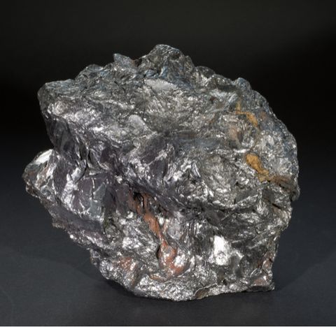 Colour photograph. A rocky form with an uneven, shiny silver surface is photographed sitting on a dark tabletop, against a black backdrop. Small traces of pink and beige can just be seen peeking through the silver.